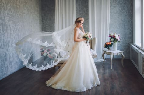 Panna młoda w eleganckiej, koronkowej sukni ślubnej z welonem, trzymająca bukiet kwiatów, stojąca w stylowo urządzonej sali weselnej.