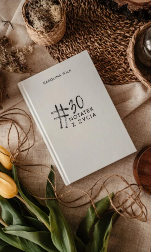 Książka #30 Notatek z Życia Karolina Wilk na tle naturalnych dekoracji z kwiatów i wikliny.